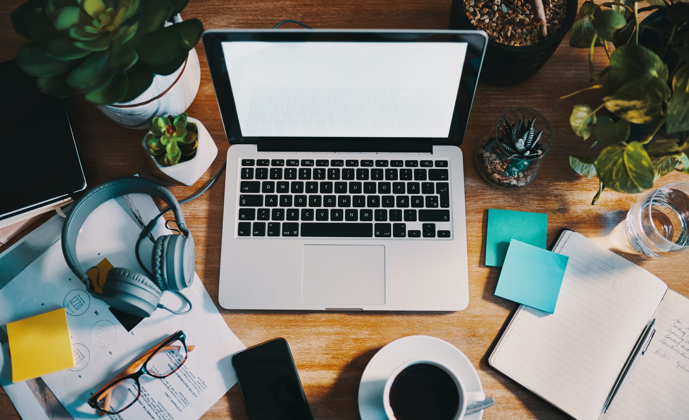 Overhead view of a desk with a laptop, plants, headphones, coffee, and notebook.