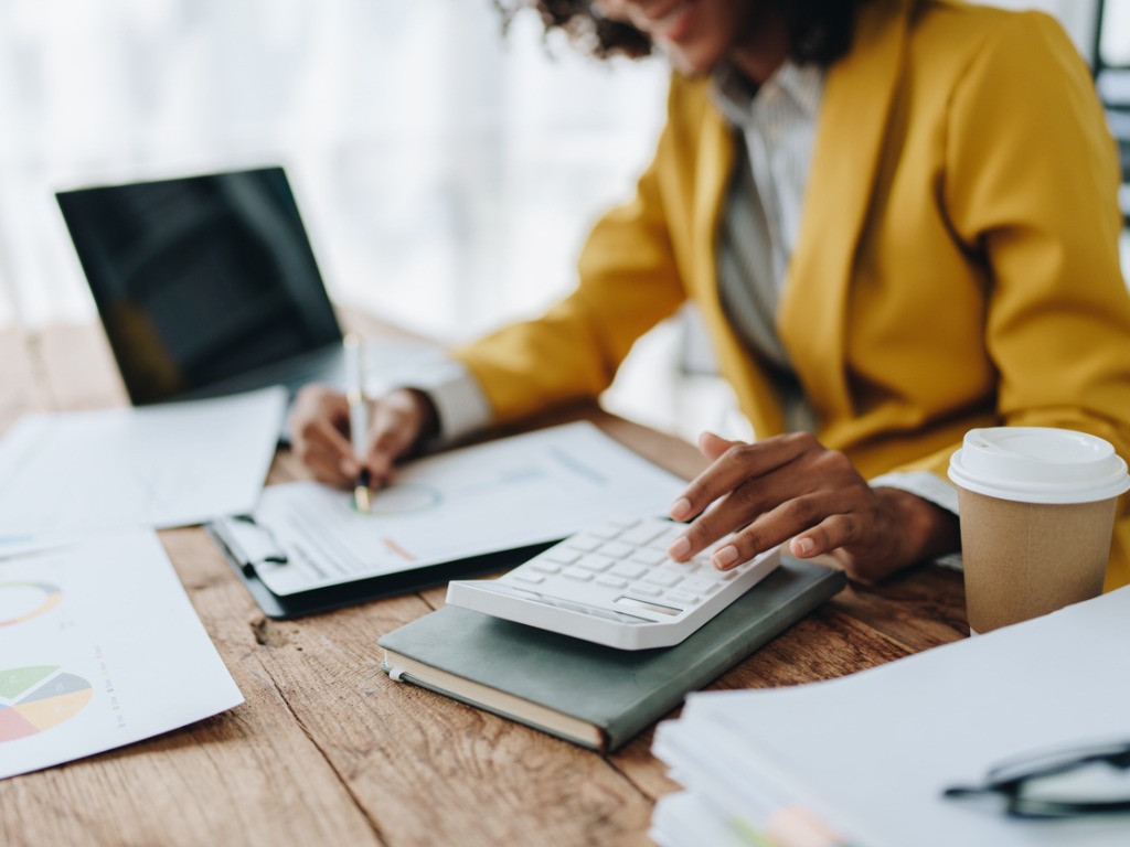 Woman in yellow blazer using calculator at desk with laptop and coffee.